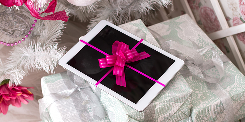 Beli tablet ukrašen roze mašnicom na gomili upakovanih novogodišnjih poklona ispod jelke