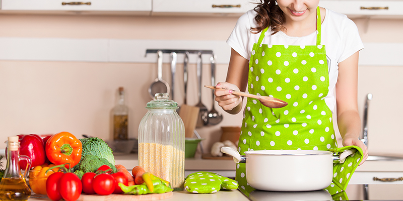 Devojkaza zelenom keceljom na bele tufne u kuhinji isprobava ručak koji se krčka na šporetu