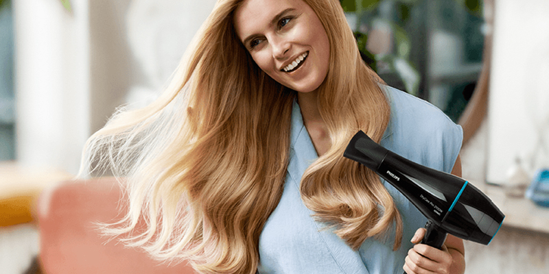 Devojka sa dugom plavom kosom sređuje frizuru profesionalnim Philips fenom za kosu crne boje.