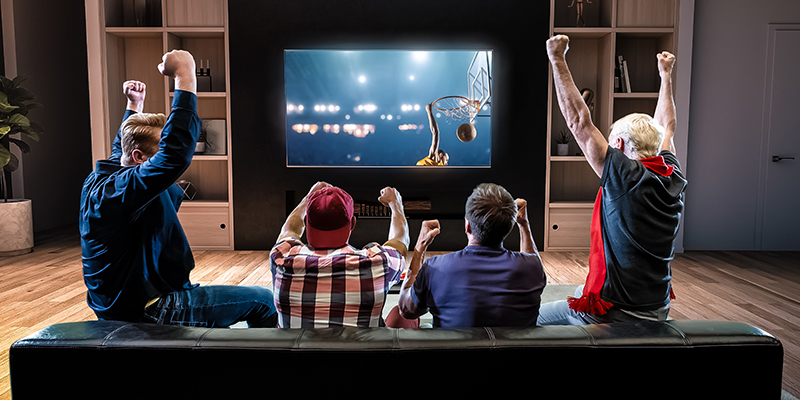 Četiri prijatelja sede na kauču u dnevnoj sobi, gledaju košarkašku utakmicu i navijaju
