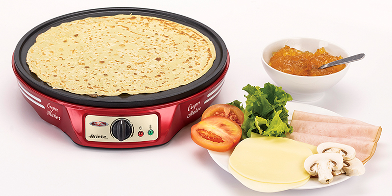 Ariete crveni aparat za palačinke na kom se peče palačinka, pored je tanjir sa suhomesnatim proizvodima i povrćem