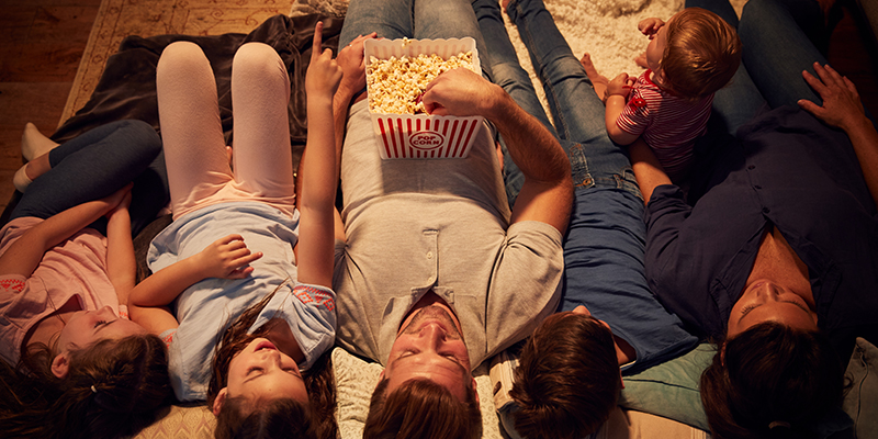 Šestočlana porodica leži na udobnom belom tepihu na kome su jastuci, jedu kokice i uživaju u zajedničkom gledanju televizije