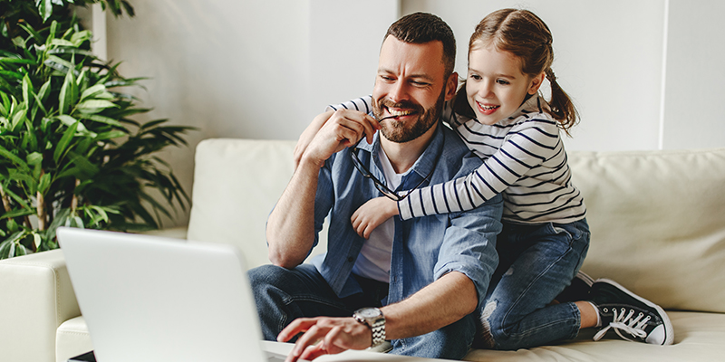 Nasmejani otac i njegova ćerkica sede na sofi - otac radi na laptopu, a ona ga je zagrlila