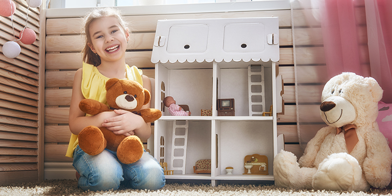 Nasmejana devojčica drži igračku medu, a iza nje se nalazi kućica za lutke