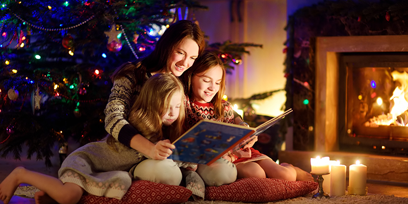 Majka sa dve ćerkice sedi na jastucima i tepihu ispred kamina u prostoriji sa novogodišnjom dekoracijom i čita im knjigu