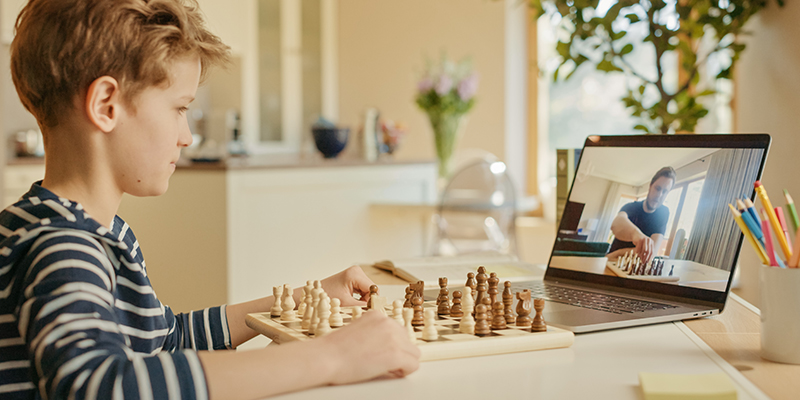 Dečak plave kose uči kako se igra šah koristeći online tutorijale na laptopu. Ispred njega je tabla sa figurama na kojoj pokušava da ponavlja korake na osnovu online instrukcija