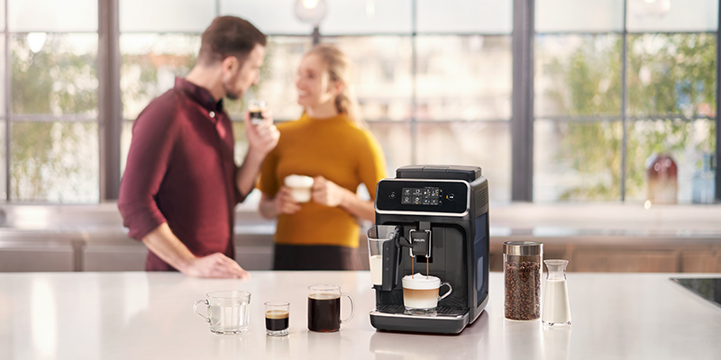 U prvom planu aparat za espresso LatteGo sa napravljenim različitim vrstama kafe na pultu, a u pozadini se zamućeno vide dečko i devojka koji drže u ruci šoljice sa kafama i ćaskaju
