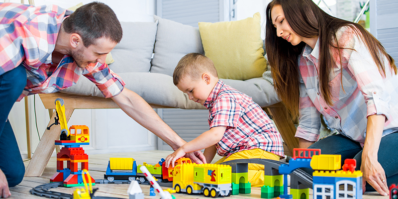 Muž i žena u kariranim košuljama i farmerkama igraju se sa detetom na podu. Za igru koriste mnogobrojne LEGO kockice. Tu su kamion, dizalica, pruga sa šinama, most i još mnoge druge konstrukcije od kockica. Pored njih je kauč sa sivo-žutim jastucima
