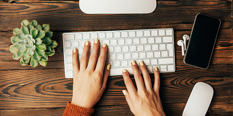 Devojka sa nalakiranim noktima koristi bežičnu belu tastaturu. Pored nje nalazi se cveće, bežične slušalice, miš i mobilni telefon