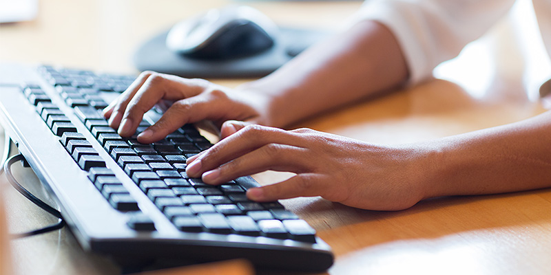 Mlada poslovna žena sedi za radnim stolom, radi na računaru koristeći standardnu, tzv. mejnstrim tastaturu
