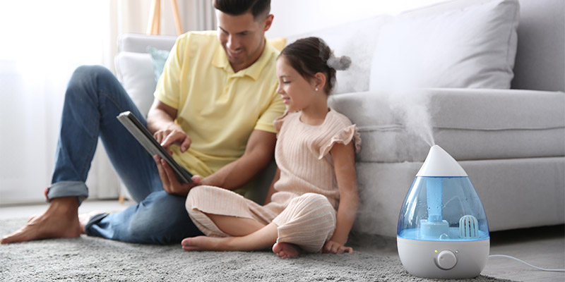 Otac i ćerka sede na tepihu ispred kauča bele boje i čitaju knjigu. Pored njih je prečišćivač vazduha koji donosi svežinu i čist vazduh u prostor u kome borave