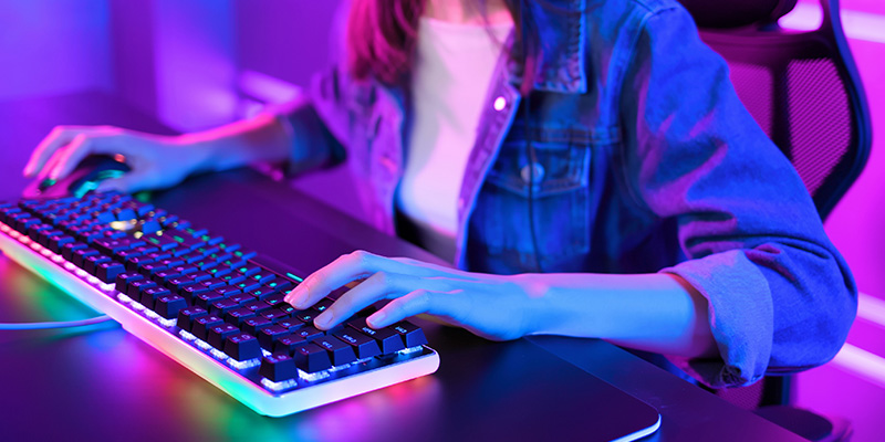 Tinejdžerka u teksas jakni i beloj majici igra igrice koristeći mehaničku tastaturu sa osvetljenjem.