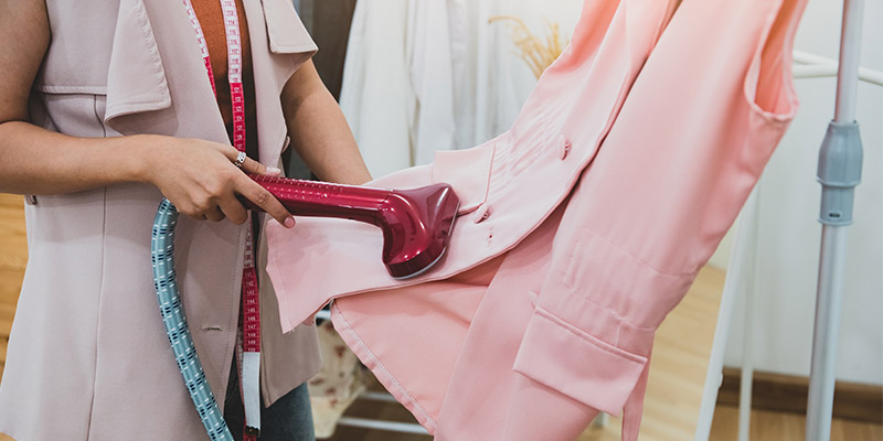 Krojačica u roze prsluku sa okačenim metrom oko vrata pegla pomoću vertikalne pegle drugi prsluk roze boje koji je okačen na držaču