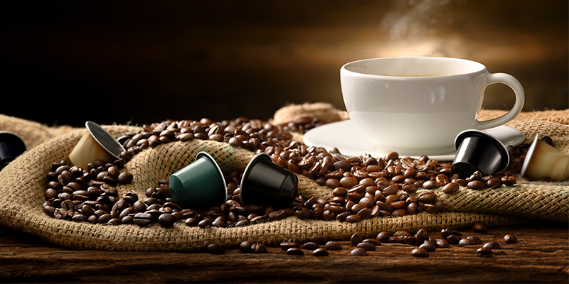 Na starinskom džaku braon boje prosuta su zrna kafe i nekoliko kapsula za kafu. Pored njih tu je I šolja sa tanjirićem iz koje se osete mirisi tek skuvane kafe