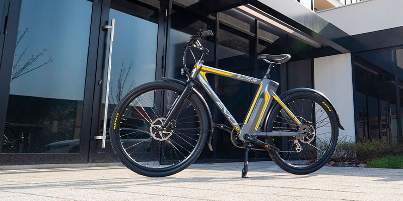 E prime električni bicikl žuto-sive boje parkiran je ispred ulaza u modernu zgradu