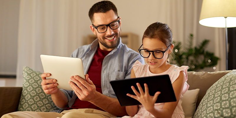 Otac i ćerka uživaju u korišćenju digitalnih uređaja uz naočare za zaštitu od plavog svetla sa ekrana