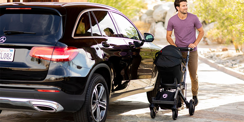 Otac moderno odeven prolazi pored parkiranih kola i vodi dete u šetnju Orbit baby kolicima crne boje u planinskim predelima