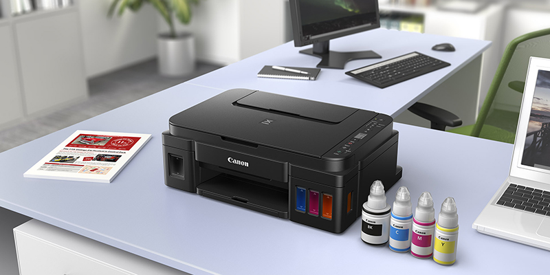 Canon Pixma G3411 štampač i mastilo u četiri boje smešteni su na radni sto pored monitora, tastature i laptopa