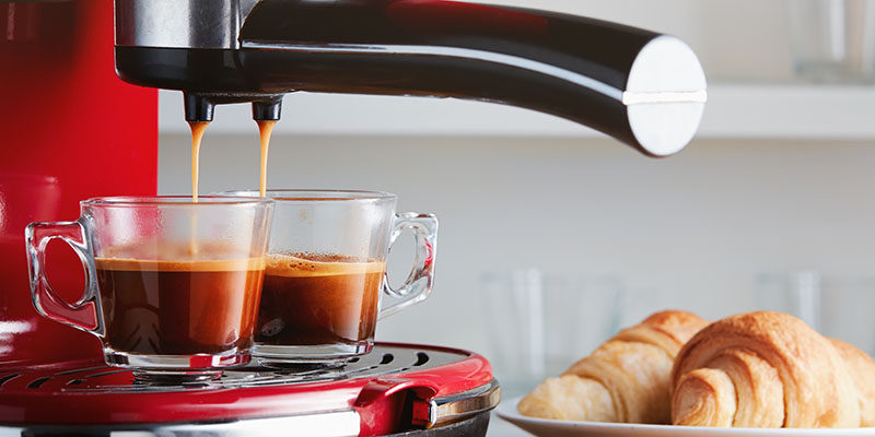 Crveni aparat za espresso koji upravo priprema kafu za dve šoljice, a pored je tanjir sa kroasanima