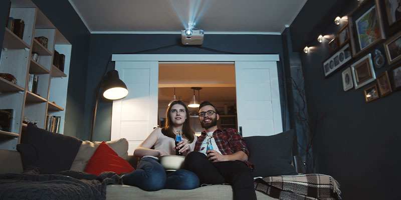 Momak i devojka sede na kauču i gledaju film koristeći projektor