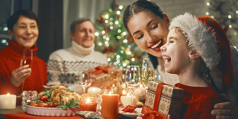 Nasmejana porodica sedi za trpezom i razmenjuju novogodišnje poklone