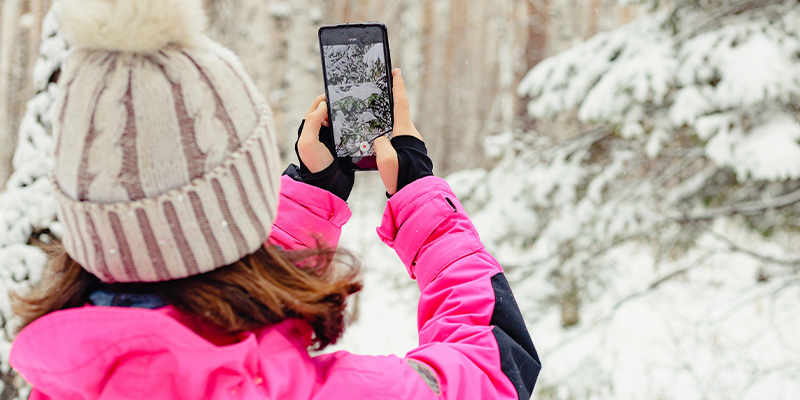 Devojka u roze zimskoj jakni i sa kapom drži telefon i slika zimski prizor sa snegom