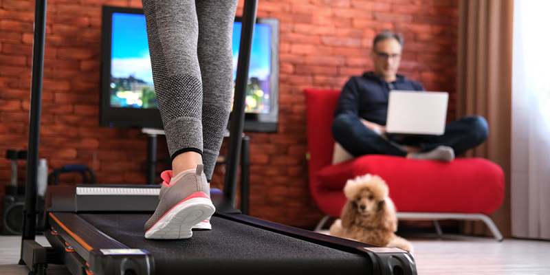 Žena vežba na traci za trčanje u dnevnoj sobi, u uglu na fotelji sedi muž i radi za računarom, a na podu je kuče